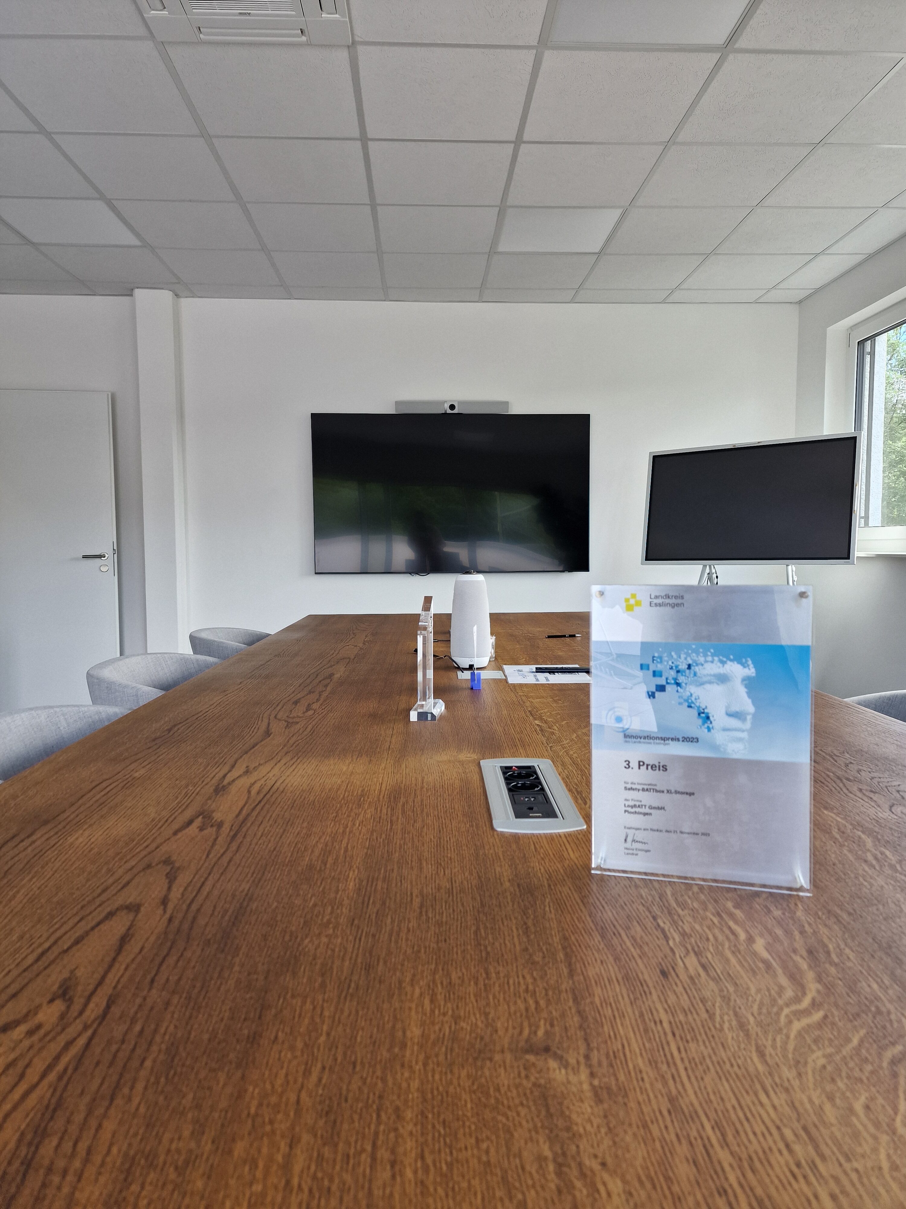 Moderner Besprechungsraum mit großem Holztisch, Bildschirm und grauen Stühlen. Helles Ambiente.